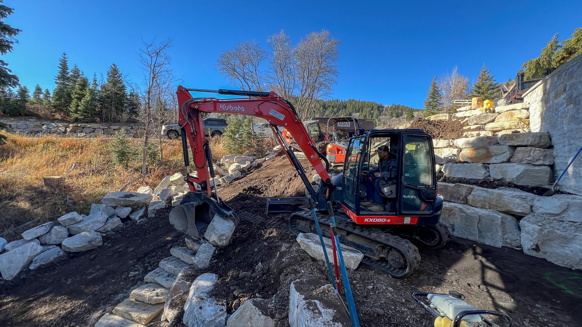 Excavator building stone retaining wall in forested area.