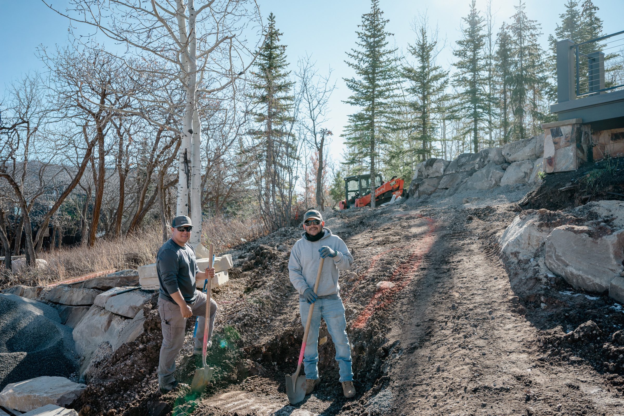 Two men digging ground near tree line and machinery.
